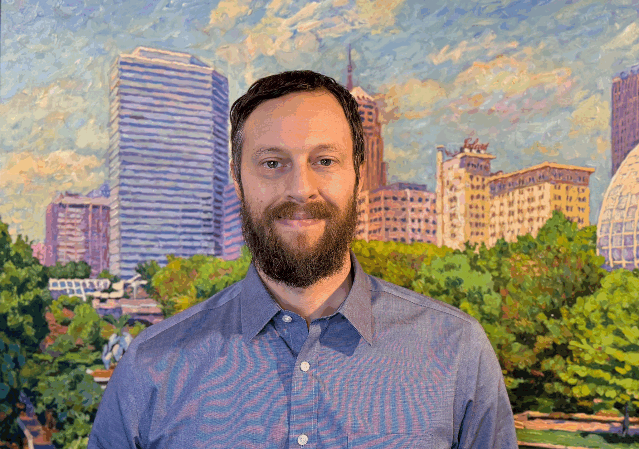 Bearded man in a blue shirt smiling in front of a cityscape painting of Oklahoma City.