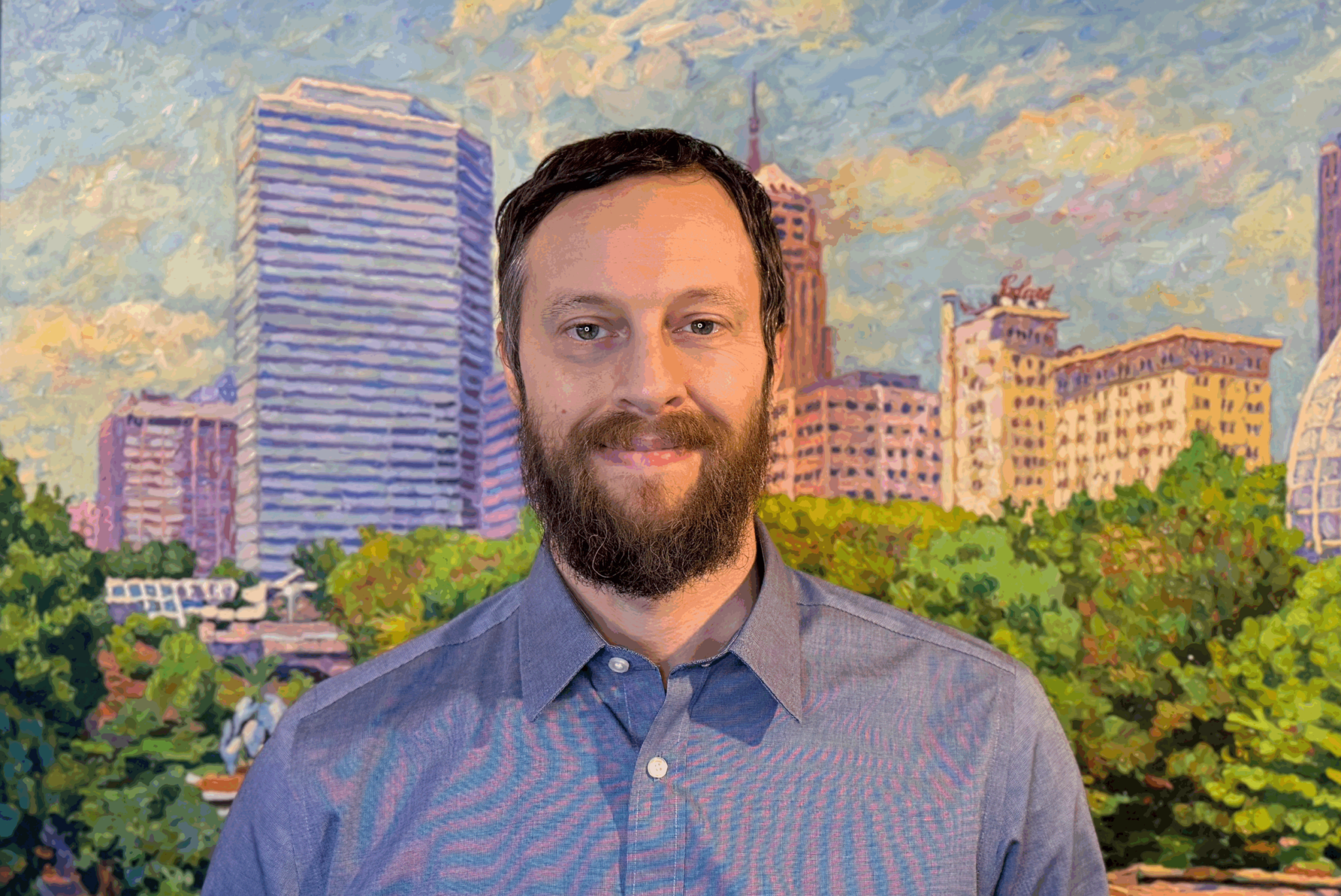 Bearded man in a blue shirt smiling in front of a cityscape painting of Oklahoma City.