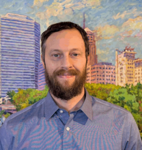 Man with beard in blue shirt in front of a painting of the Oklahoma City skyline.
