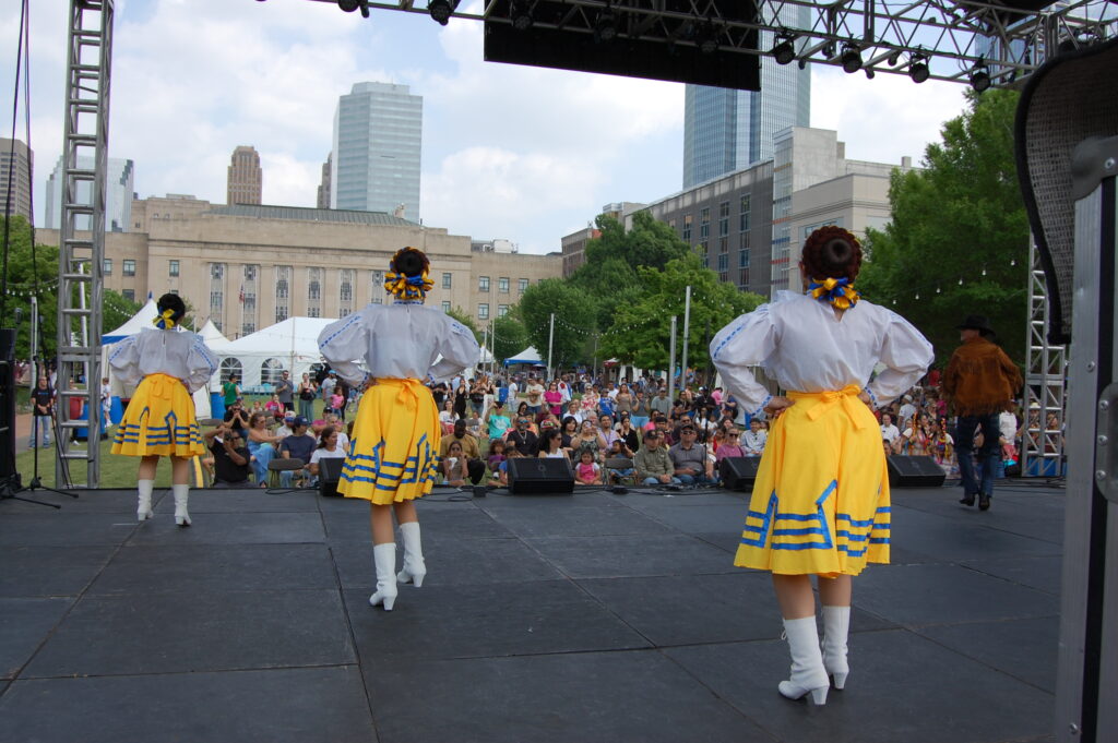 Reverse view of dancers in bright costume on stage facing an outdoor festival crowd.