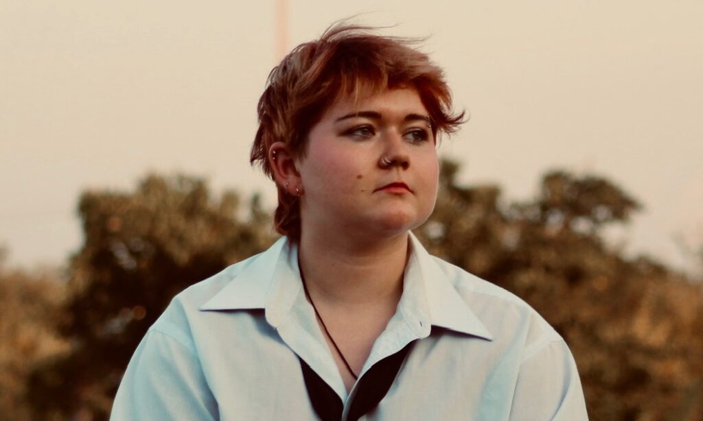 Portrait of Teaching Artist Astrid Moan, a non-binary person, seated in front of treeline at sunset.